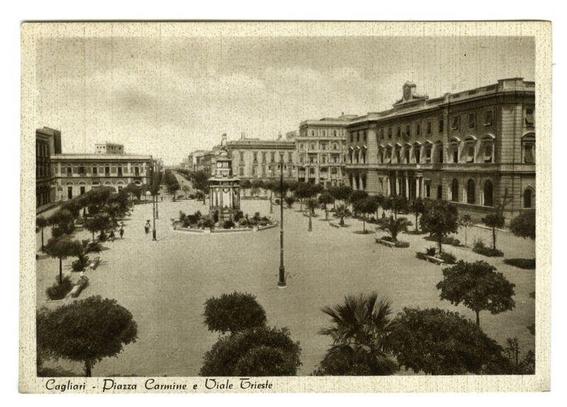 The image depicts a historical black-and-white photograph of Piazza Carmine in Cagliari, Italy. The perspective is from an elevated vantage point looking down onto the square and surrounding streets. Prominent features include neoclassical buildings with multiple stories and rows of windows along both sides of the street leading into the plaza.

In the center of the piazza stands a prominent column structure that appears to be a fountain or monument, surrounded by palm trees planted in circular beds at regular intervals. A few individuals can be seen walking on the wide streets, which are lined with lampposts and more greenery including shrubs and larger bushes. The atmosphere suggests an early 20th-century setting based on architectural styles and clothing of people visible.

The photograph has a caption that reads "Cagliari - Piazza Carmine e Viale Trieste," indicating the location as Cagliari, with specific mention to these two landmarks. There is some wear and discoloration on the physical print or digital representation, which adds texture but does not detract from the clarity of the historical features within it.

For a more detailed description including information about this photograph such as how it might be used in different contexts (e.g., academic research, tourism promotion) please refer to external resources.