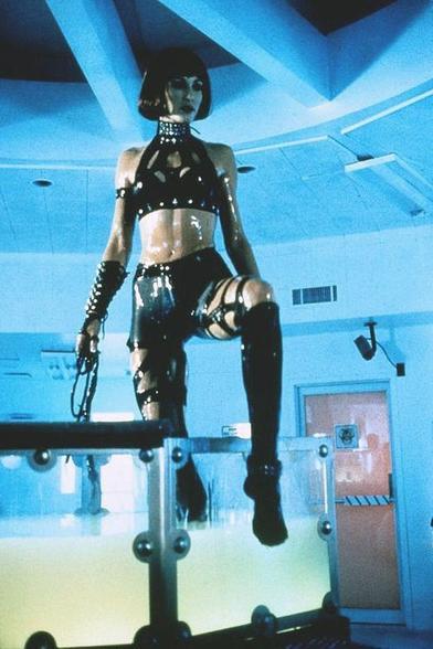 A still from the movie Virtual Combat (1995) featuring the character Greta (played by Dawn Ann Billings). She stands authoritatively on a glass structure in a blue-lit room, wearing a glossy black PVC outfit with a studded choker and holding a whip in her right hand.