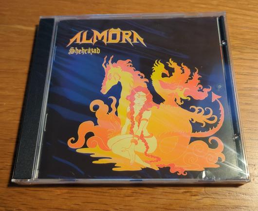 An cd album is on a wooden table. It says almora on it and there is a flaming horse with a ancient women figure sitting on the ground in front of it.