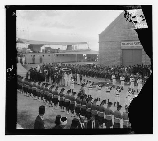 A black and white photograph capturing a formal military ceremony or parade. The image shows rows of soldiers in uniform, some holding flags, standing at attention while others march along the ground. A building with "TRANSIT SHEED" written on it is visible to one side, suggesting an industrial setting such as a depot or terminal related to transport services. Spectators are present around the perimeter, observing the event. The photo appears aged and has markings indicating that it was part of American Colony Photographic Department collection in Jerusalem from 1933.