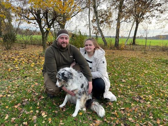 Zwei Personen mit einem schwarz-weißen Schäferhund im Freien, Herbstlandschaft mit gelben Bäumen im Hintergrund.
