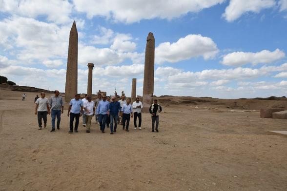 Gruppo di archeologi che camminano tra obelischi e rovine nel sito di San el-Hagar, Egitto, con cielo nuvoloso sullo sfondo.
