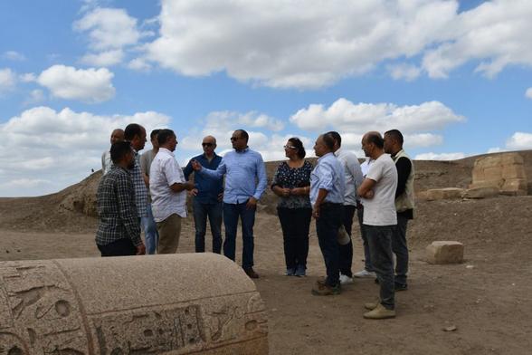 Un gruppo di archeologi e studiosi discute presso un sito archeologico nel Delta orientale dell'Egitto, con rovine antiche visibili sullo sfondo e un cielo parzialmente nuvoloso.