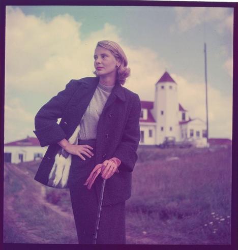 a woman in a coat with red gloves looks over her shoulder as she stands outside near some houses.  the photo is tinted purple and has an old feel to it.