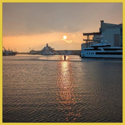 In a broad quiet water stands a mooring post in full backlight. You also see the wintersun. To the left and the right there are large luxury yachts and af fishing boat.