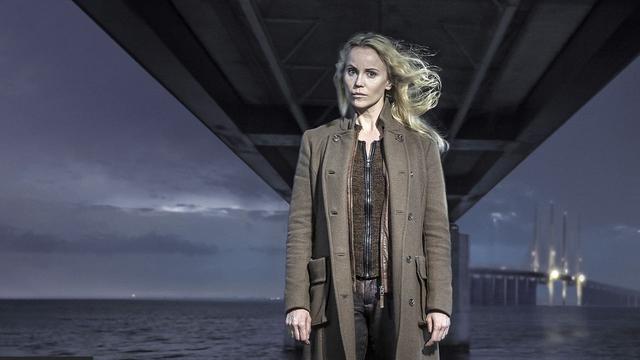 Sofia Helin mit langem, offenem, blonden Haar und braunem Mantel steht unter der Öresundbrücke über Wasser, im Hintergrund beleuchtete Brückenpfeiler. (Bild: © 2013 Filmlance International AB/Carolina Romare )
