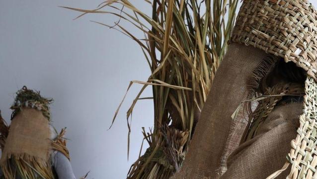 Image: photo – we see a sheaf of grass of some sort quite close to the camera, and it seems that it may have provided the material for two woven strips that we see towards top right and right in the photo; there seems to be more sheafery hidden behind burlap or equivalent, and farther away we may be seeing someone wearing this gear, along with some grass and stones, maybe, on their head; a grey wall behind • “^ is a collective based in Manorhamilton, Leitrim. Our five members are Tara Baoth Mooney, Shane Finan, James Kelly, Laura McMorrow, and Sonya Swarte. We collectively occupy an artist-led studio and experimental space in Manorhamilton, opened in September 2022. Over the past three years, we have led events in north Leitrim in response to environmental grief, care, and tending to the land.  For this exhibition-environment, we explore meadows, untrodden ground, and forgotten growth in north Leitrim. In these liminal spaces, a succession of plants create worlds upon worlds, each…”