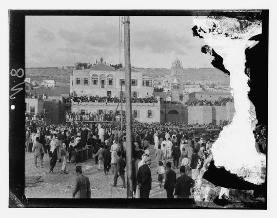 The image depicts a bustling public gathering, likely during the 1930s based on historical context. It is a black and white photograph with high contrast, showcasing numerous individuals walking or standing in what appears to be an urban setting.

In the background stands prominent architecture suggestive of Middle Eastern design, including buildings with arched windows, domes, and spires that are characteristic of Islamic-inspired structures. One such building features multiple archways leading into a larger complex, while another has a clock tower-like structure, indicating it may serve as a significant public or administrative edifice.

The crowd is diverse in terms of gender and attire, suggesting the event holds cultural significance for various segments of society. Men are dressed in traditional Middle Eastern garments such as long tunics (jubbas), headscarves known as ghutras or keffiyehs, and some wear hats like fezzes or turbans.

The photo has a large white overexposed section on the right side, obscuring part of the scene. This stark contrast between light and dark gives the image an almost surreal quality, emphasizing texture and form while diminishing detail in shadowed areas.

There's also text at the top left corner indicating "1938" with additional writing that is not entirely legible due to photo condition or resolution limitations.

Overall, this photograph captures a moment of historical significance,  [...]