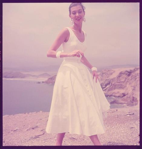 The image shows a woman in an elegant white dress standing on rocky terrain with the ocean and distant hills visible in the background. She is smiling warmly, wearing large hoop earrings and multiple bracelets or bangles on her wrists. The photo has a vintage aesthetic typical of mid-20th-century fashion photography, characterized by pastel tones that give it a warm, nostalgic feel.

Additional information indicates this photograph was taken for "Fashion" magazine in 1951 as part of the Toni Frissell Photograph Collection, which features images of celebrities and socialites from America during that era.