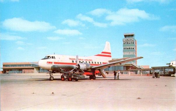 A vintage postcard-style scene shows a Trans World Airlines (TWA) propeller aircraft parked on a sunny airport ramp. The plane is white with bold red stripes and the classic TWA logo along its fuselage. Its twin propellers sit still while a small service vehicle is positioned beneath the nose. A few ground crew members stand nearby, appearing relaxed as they work or talk. Behind the aircraft is a low, modernist terminal building with long horizontal windows. Rising above it is a tall control tower with large block letters spelling “WICHITA” near the top. The sky is bright blue, dotted with soft clouds, giving the whole scene a clean, optimistic mid-century feel.