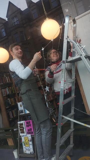 booksellers Jack and Olivia on a ladder in the window of the Edinburgh Bookshop, untangling a line of fairy lights