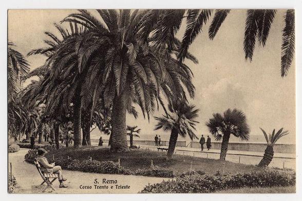 The image is a sepia-toned photograph depicting an outdoor scene. It appears to be from the early 20th century, judging by its style and quality. The location identified on the lower part of the photo reads "S. Remo Corso Trento e Trieste," indicating it was taken in Sanremo along the Corso Trento E Trieste.

The scene shows a promenade lined with palm trees and other tropical vegetation, suggesting a coastal or seaside setting. There is an individual seated on a folding chair under one of these palms, seemingly relaxing while possibly reading something, as they are holding what appears to be a book or magazine in their lap.

In the background, there's a view extending towards the sea with several people walking along the promenade, enjoying leisurely strolls. A few more individuals can also be seen standing near what looks like a small gazebo-like structure on the beachfront. The sky is overcast, and there are no bright colors due to the monochromatic nature of the photograph.

The image has a serene vibe, emphasizing relaxation and the enjoyment of outdoor leisure activities in a picturesque environment.
