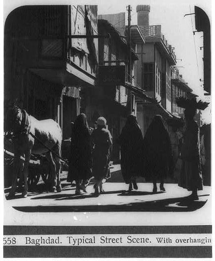 The image depicts a bustling street scene from Baghdad in 1932, captured by the Matson Archive. It features several individuals dressed in traditional attire walking along a narrow urban pathway lined with buildings and overhanging balconies that cast shadows on the pavement below.

In the foreground, we see three figures wearing cloaks or long robes indicative of Islamic dress code, suggesting this scene is set during a time when such garments were common. Their heads are mostly covered except for their faces, emphasizing modesty as part of cultural attire in many regions of the Middle East at that period.

On the left side of the image stands a horse-drawn carriage or cart being operated by an individual not fully visible within this frame; only parts of their body and possibly reins or harnesses can be seen. This mode of transportation adds to the historical context, suggesting pre-motorized travel was still in use during that era.

The architecture includes buildings with multiple levels featuring balconies supported by ornate railings which could suggest European influences on local architectural styles typical for Baghdad's diverse cultural amalgamation. The contrast between light and shadow is striking; it highlights textures like the wooden balustrades, rough plaster walls, or stone paths indicative of an older city.

The text at the bottom indicates that this image represents "Baghdad. Typical Street Scene. With [...]