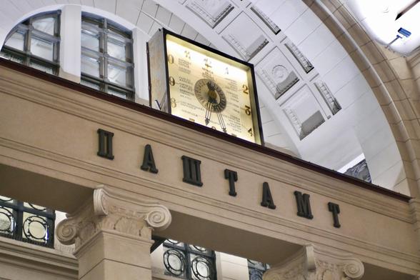 A close up shot of the square "World clock" with the names of major world cities written in Belarusian on the watchface, and the word ПАШТАМТ (Pashtamt, "Post office" in Belarusian) beneath it on the façade of the Main postal office in Minsk, Belarus, on an October evening in 2025.