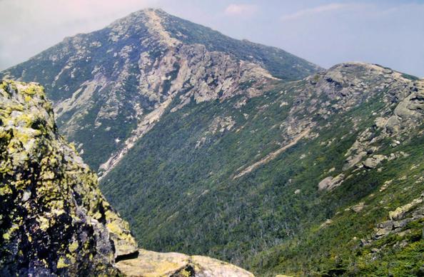 Beneath a hazy light blue sky with several small hazy clouds to our right and a larger one to our left, we are standing next to a large light gray granite outcrop covered with light and dark green lichen. This outcrop frames the bottom lefthand corner. A rocky alpine ridge and peak dominates our view. It is made of the same light gray granite as the outcrop, with bands of very low green alpine vegetation.