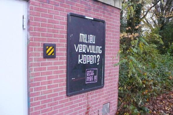 stenen muur met witte deur links en struiken rechts
reclame frame met zwart gemaakte poster met witte letters
en daar onder oze letters omlijnd
op het muurtje nog wat roosters