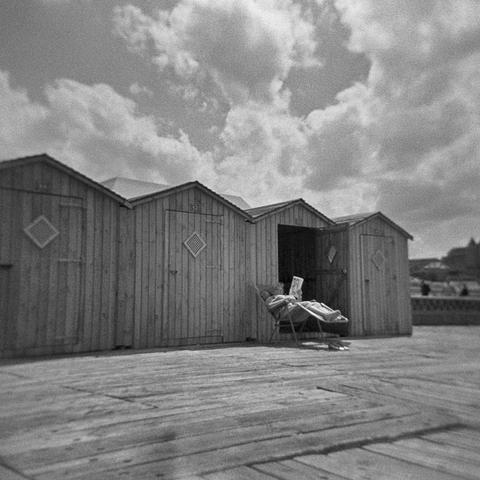 photo en noir et blanc avec du grain qui montre des cabines de plage en bois avec l'une à la porte ouverte et juste devant une personne dans un transat en train de lire un journal.