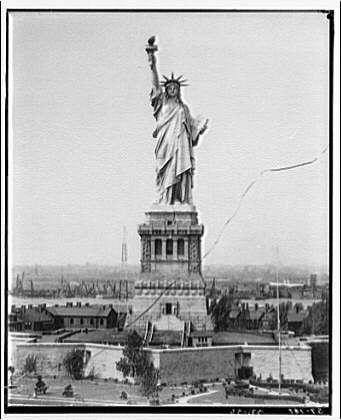 The image depicts the iconic Statue of Liberty standing tall against a clear sky, viewed from an aerial perspective. The statue is captured in black and white, emphasizing its architectural details and grandeur. It holds up a torch aloft with one hand while clasping something resembling a tablet or book in the other.

In the background, we see an expansive view that includes parts of New York Harbor along with some buildings on shorelines visible at various distances behind it. The image appears to be from around 1920-1950 based on its visual style and quality; however, without additional context or metadata attached to this particular file, determining the exact date remains speculative.

The photograph captures a moment of historical significance in American history when the Statue of Liberty was being constructed. This landmark is one of New York City's most recognizable symbols, representing freedom and democracy since it was dedicated on October 28th, 1886.