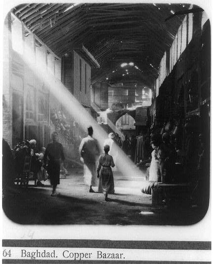 A black and white photograph depicting a bustling market scene within the Copper Bazaar in Baghdad. Sunlight streams through an arched entryway, casting long shadows across the alley as people walk towards or away from it. A woman is prominently featured walking with her child on one side of this shadowed beam of sunlight while others are seen further down the lane engaged in various activities such as shopping and conversing. The photograph carries a historical quality indicative of early 20th-century Middle Eastern life, reflecting an era when street markets were vital hubs for commerce and social interaction.