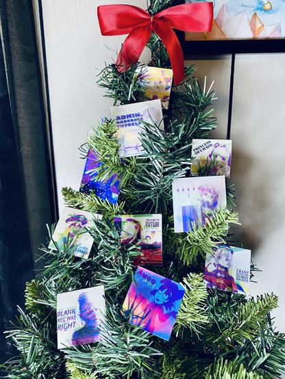 A small decorated Christmas tree featuring various colorful stickers with artistic designs and phrases. The tree is topped with a bright red bow and adorned with stickers displaying messages related to identity and creativity. The background suggests a cozy indoor setting.