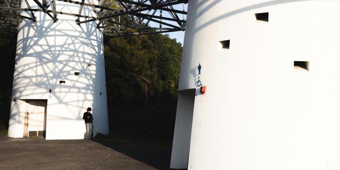 White cylindrical structures with geometric shadows cast by an overhead metal framework. A man stands near one structure with a wheelchair-accessible symbol visible. Sunlit trees in the background create contrast with the architectural elements.