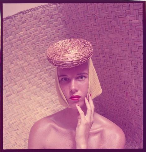 The image depicts a person with pale skin and red lips, wearing what appears to be an eyepatch or bandage on their face. They are also adorned in a hat that is brownish-beige in color, which has been styled into a rounded shape atop the head. The individual's fingers have bright pink nail polish, contrasting against the muted tones of their skin and clothing.

The background consists of what looks like tightly woven wicker or rattan material, suggesting an outdoor setting such as a garden chair or bench. This creates a casual yet stylish ambiance for the portrait.

A notable feature is that the photo appears to have been altered with a pinkish-purple hue applied over it, giving the image a vintage or stylized look reminiscent of fashion photography from past decades.