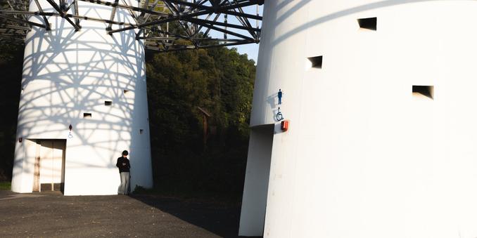 White cylindrical structures with geometric shadows cast by an overhead metal framework. A man stands near one structure with a wheelchair-accessible symbol visible. Sunlit trees in the background create contrast with the architectural elements.