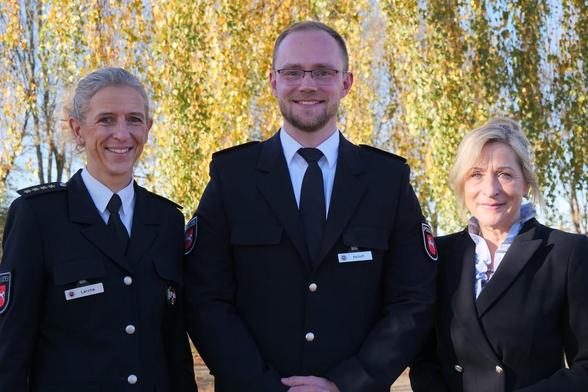 Drei Personen in dunkelblauen Uniformen mit Abzeichen stehen lächelnd vor herbstlichem Hintergrund.