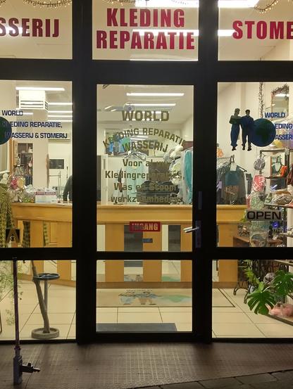 Kijk door de winkelruimte van World Kledingreparatie in Wassenaar, met een door tl-buizen verlichte ruimye met een balie en naaispullen. Op de ruit staan de teksten "Wasserij", "Kledingreparatie" en "Stomerij" in rode kapitalen. ¦ Look through the shop window of World Kledingreparatie in Wassenaar, with a fluorescent-lit space containing a counter and sewing supplies. The window displays "Laundry," "Clothing Repair," and "Dry Cleaning" in red capital letters in Dutch.