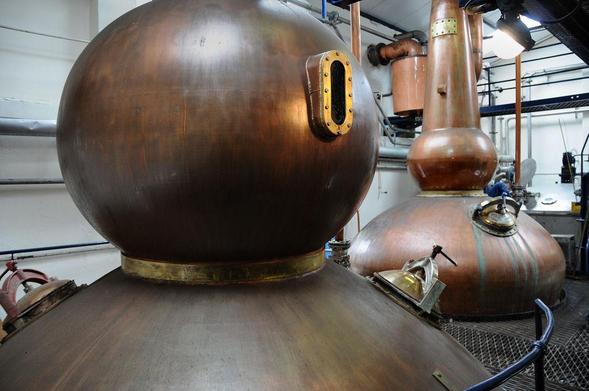 The highly distinctive stills at Pulteney Distillery, Wick. The image shows two stills, with the one in the foreground having a large spherical upper chamber and a viewing window that looks like something from a science fiction comic. The copper of the stills is quite dull and stained.