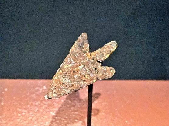A corroded metal arrowhead displayed upright on a thin stand. The triangular point is heavily weathered and rough, with two short side barbs extending from the base. Its surface shows a mottled texture of rust and mineral deposits, giving it an uneven, pitted appearance