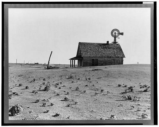 The image shows a black and white photograph of an old, weathered farm house in the middle of a barren field. The structure appears to be made from wooden planks with visible signs of decay and damage. It has a gabled roof that slopes down on all sides, which is covered with shingles showing significant wear. A windmill sits atop one corner of the building, suggesting reliance on wind power for pumping water or generating electricity in an era before modern electrical systems were widespread.

The surrounding area looks desolate; it's hard to tell what kind of vegetation remains due to the black and white nature of the photo but there are patches that look like dry earth with sparse growth. The field extends far into the background, merging with a hazy horizon under an overcast sky which adds to the overall bleak atmosphere.

The setting suggests this could be in rural America during one of its more challenging periods, likely reflecting on the Dust Bowl era where farming communities faced droughts and severe weather conditions leading to soil erosion. The photo is credited as being part of "Dust Bowl farm" from Coldwater District, north of Dalhart, Texas. It notes that this house appears occupied while most others in the district are abandoned.

The photograph seems to capture a moment frozen in time, telling a story of resilience and struggle against environmental forces beyond human control during what were likely dif [...]