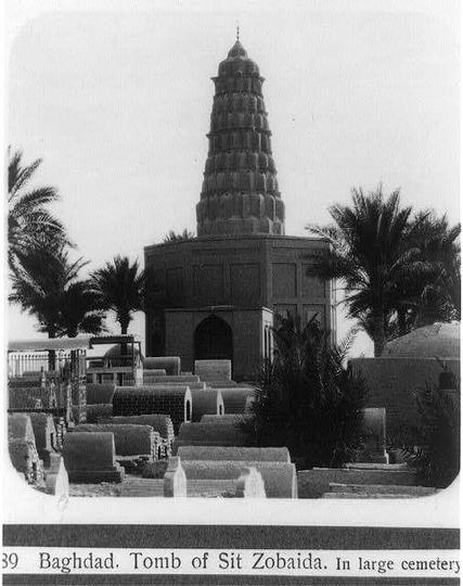 The image depicts the tomb of Sit Zobaida, located in a large cemetery near Baghdad. The structure is characterized by its tall, tiered tower that resembles an Asian pagoda and stands prominently amidst several grave markers. Palm trees are scattered around the site, enhancing the serene ambiance typical for burial grounds or memorials from this era. In the foreground, rows of gravestones can be seen, suggesting a historical context likely dating back to early 20th-century Middle Eastern architecture and cemetery practices. The black-and-white nature of the photograph emphasizes its vintage quality, capturing an architectural landmark with cultural significance in Baghdad's history.