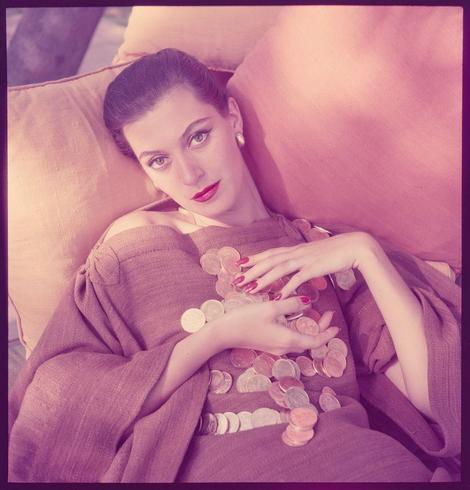 A portrait of a woman reclining on cushions, wearing an ornate dress with coins attached to it. The image has a pinkish hue and appears to be vintage in style. Notable features include the woman's confident gaze and red lipstick; she is also holding or surrounded by various coins which may symbolize wealth or value