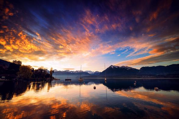 Diese Weitwinkelaufnahme fängt die majestätische Schönheit eines alpinen Sees bei Sonnenaufgang ein. Ein Himmel voller dramatischer orange- und purpurfarbener Wolken spiegelt sich im ruhigen Wasser. Die schneebedeckten Gipfel der Berner Alpen bilden eine beeindruckende Kulisse für die friedlich ankernden Segelboote.