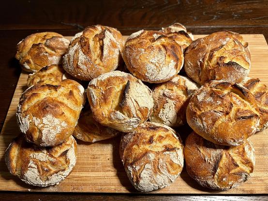 Auf einem dunklen Holztisch liegt ein Holzbrett. Darauf liegen zahlreiche frisch gebackene Brötchen mit rustikalem Ausbund.