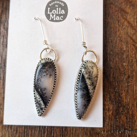 Earrings on a LollaMac display card, featuring dark elongated teardrop shapes with white and gold highlights, on a silver bezel mounted on a ring from an earwire.
