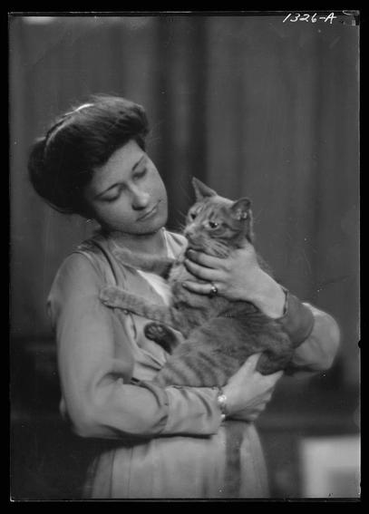 The image is a black and white portrait photograph depicting an individual holding a cat affectionately against their chest. The person appears to be wearing a sleeveless coat or jacket with decorative buttons, while the bottom half of the attire seems to have a skirt-like design. They are adorned with jewelry such as earrings and possibly bracelets on one wrist.
The subject is tenderly cradling a tabby domestic cat in their arms, holding it close, suggesting an intimate bond between them. The setting appears indoors with neutral-toned backgrounds that do not distract from the main subjects.
There's text at the top of the image which reads "1326-A," possibly indicating a catalog or inventory number associated with this photograph.