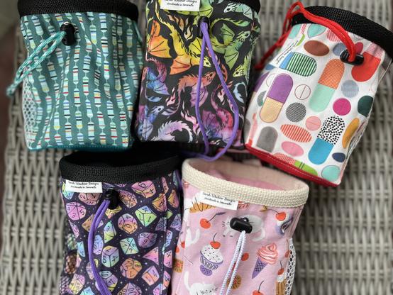 Five chalk bags on a grey wicker surface.
The top row is a teal bag with electronics resistors, a black bag with rainbow dragons, a white bag with brightly colored pills.
The bottom row is a black bag with pastel ombre ttrpg dice and a pink bag with cute kittens and desserts on it.