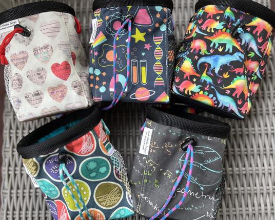 Five chalk bags on a grey wicker surface.
The top row is a cream bag with library due-date cards in heart shapes, a navy blue bag with bright pink science-emblems (DNA, test tubes, magnets, beakers, etc.), and a black bag with rainbow watercolor dinosaurs.
The bottom row is a teal bag with brightly colored petri dishes and a grey bag with chalkboard-style math equations and diagrams.