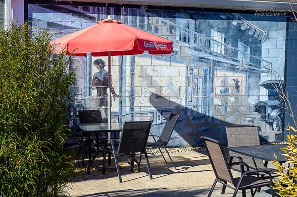 A wall painting of a street scene with 2 story buildings and a women painted in shades of blue is behind a restaurant patio with black metal chairs and tables and a red table umbrella.