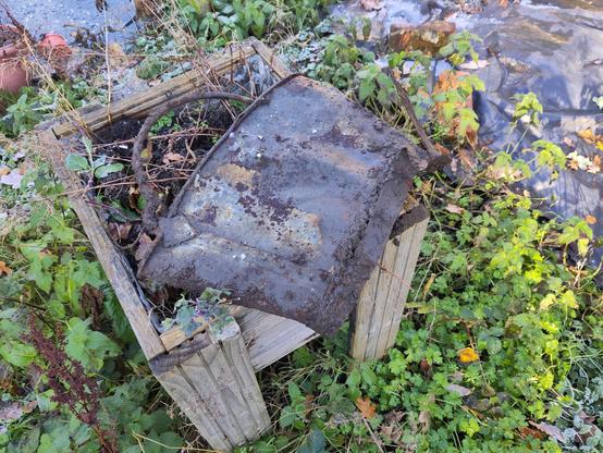 An old metal object that is flattened and discoloured after being buried under rubble. It was either a jam pan or coal scuttle. It is on a small wooden planter.