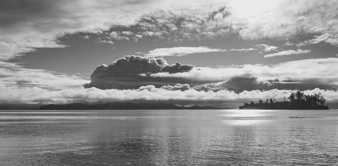 A picture taken looking across the San Francisco Bay from San Rafael towards Richmond. The Richmond Bridge just visible far right, Marin Island on the right too. A bollowing cumulus cloud in the center, dark and menacing, lighter clouds around it and a layer just above the land. Monochrome.
