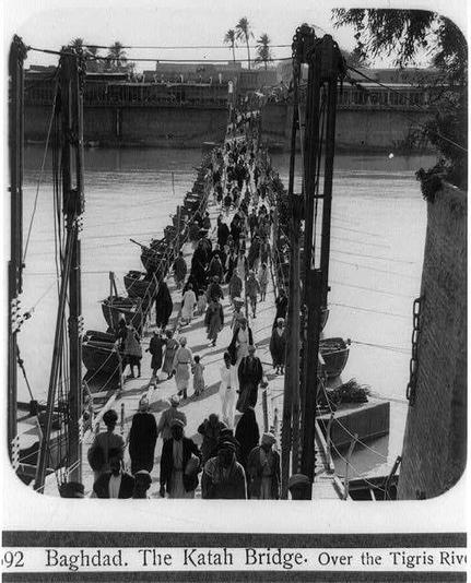 The image depicts a historic scene of people crossing the Katah Bridge in Baghdad, spanning over the Tigris River. The black and white photograph captures individuals walking across narrow wooden planks that form the bridge's path, held up by tall poles on either side which are secured to boats floating below. A dense crowd is seen traversing the makeshift structure; some people use a cane or hand support for stability as they move along.

The scene appears crowded yet orderly, suggesting it was taken during a busy period of transit across the river. The bridge seems rudimentary and temporary in construction, indicating that this may have been captured at a time when infrastructure development might not be advanced enough to construct a more permanent crossing over such a wide expanse.

In terms of background context, Baghdad's Tigris River is one of its defining natural features, centralizing the city’s historical landscape. The bridge serves as an essential pathway for movement across this significant waterway in the heart of what was once part of Mesopotamia, which includes present-day Iraq among other nations.

This photograph carries a sense of daily life and transportation challenges faced during that period, offering insights into both urban infrastructure development and social activities within Baghdad. It is notable as an artifact from 1932, shedding light on historical conditions in the region at that time.