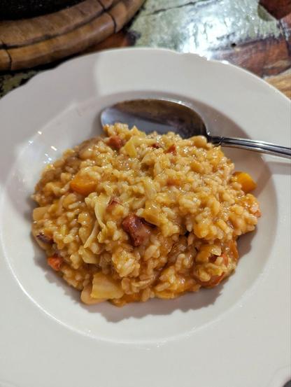 Holztisch, darauf ein weißer, tiefer Porzellanteller. Im Teller ein Reishaufen. Die rundlichen Reiskörner sind ummantelt von einer bräunlich-roten Sauce. Karotten-, Wurst-, Kohlstücke, sowie vereinzelt weiße Bohnen sind in der Masse erkennbar. Ein Esslöffel  liegt mit im Teller.