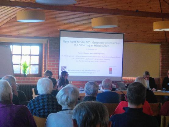 Foto aus dem Tagungsraum in der HÖB Papenburg, im Vordergrund unten Hinterköpfe der Zuhörenden, dahinter halb verdeckt die Referentinnen auf dem Podium und eine Leinwand mit Anzeige des Programms des zweiten Panels "Zukunft der Erinnerungsarbeit".