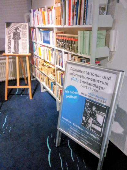 In den neuen Räumen des DIZ in der Papenburger Innenstadt: rechts im Vordergrund ein Aufsteller mit Info-Plakat zum DIZ vor einem vollen Bücherregal, links im Hintergrund eine Staffelei, auf der ein Plakat zum Thema Moorsoldaten steht.