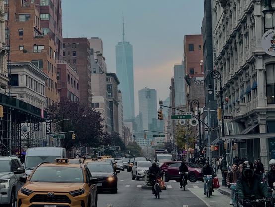 Afternoon in Manhattan. NYC’s Freedom Tower skyscraper can be seen in the distance. A yellow cab, other cars, and bicycles all jockey for space in the foreground. The scene is busy.