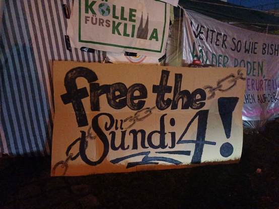 An einem Pavillon hängenen mehrere Banner, zum Beispiel "free the Sündi4!" und eine wei-grüne Fahne von "Kölle für's Klima"