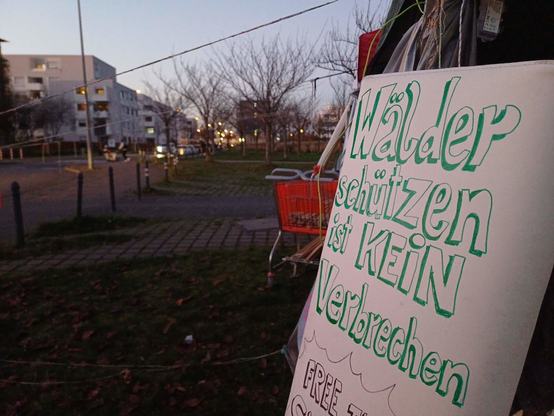 An einem Pavillon hängt ein Pappschild: "Wälder schützen ist kein Verbrechen", imHindergrund steht ein leerer Einkaufswagen am Eingang zu einem Park, weiter hinten ist das Polizeipräsidium zu sehen.